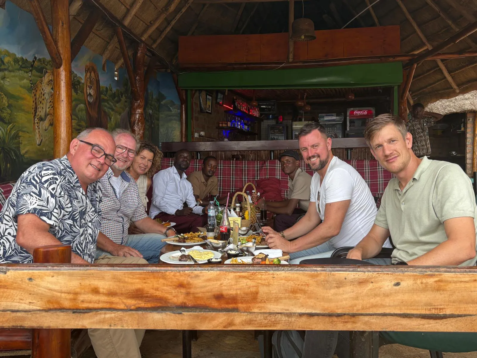 Een gezelschap rond een lange houten tafel onder een rieten dak in Oeganda